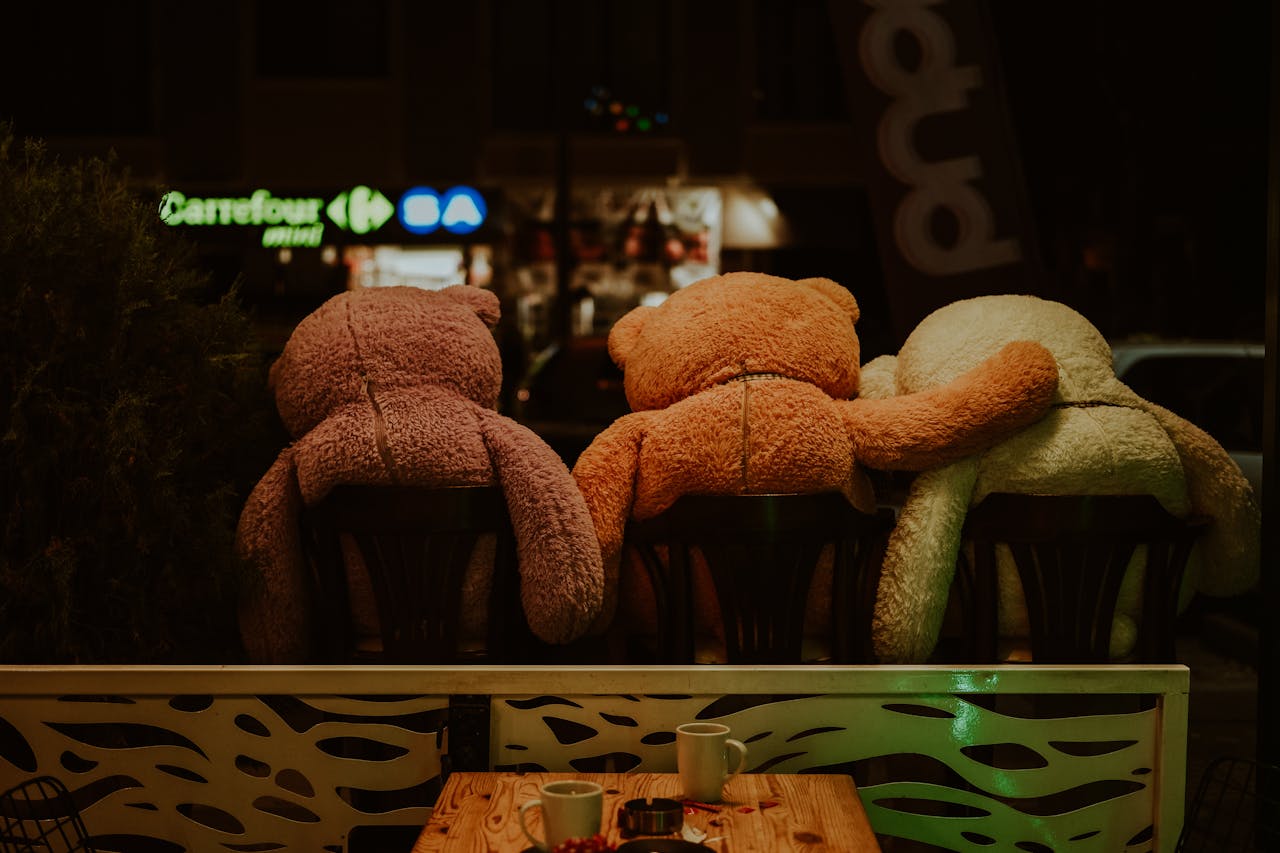 Three teddy bears sit closely on chairs at an outdoor café, symbolizing friendship and warmth.
